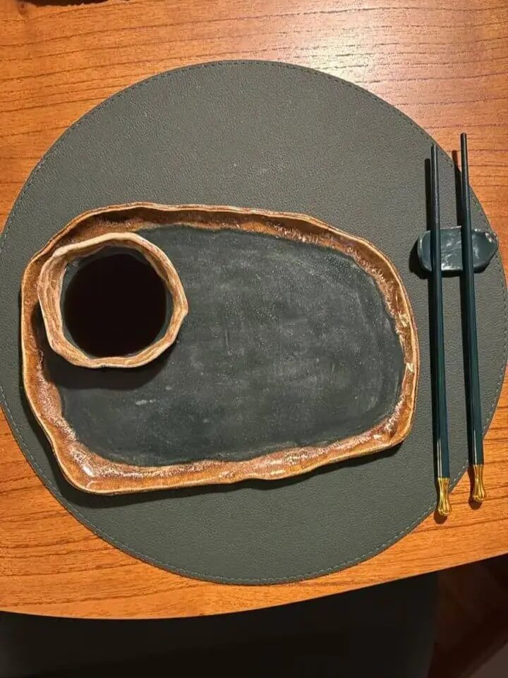 Foto de kit sushi com prato e cumbuca de shoyu, sobre a mesa, ao lado de um par de hashis.