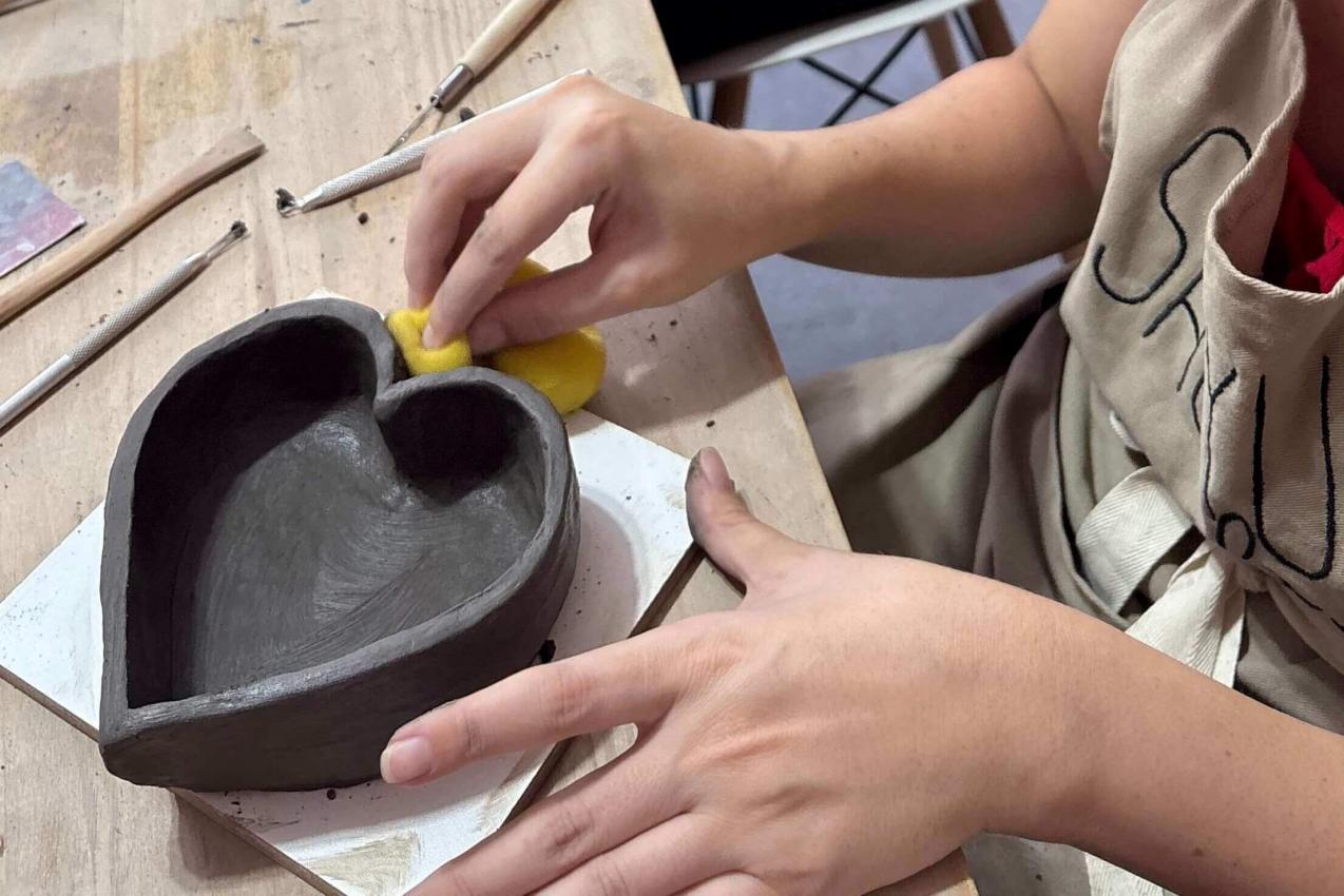 Mulher passando esponja em pote modelado à mão, em formato de coração, durante oficina de cerâmica artesanal no ateliê Shoyu Crafts.