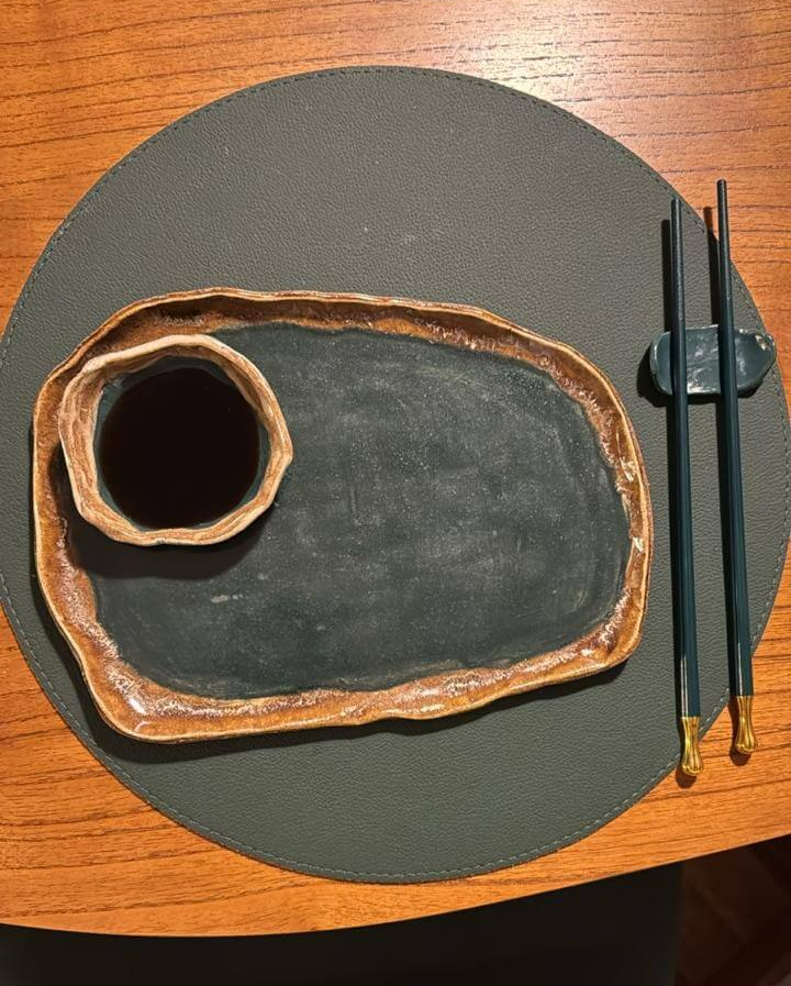 Foto de kit sushi com prato e cumbuca de shoyu, sobre a mesa, ao lado de um par de hashis.