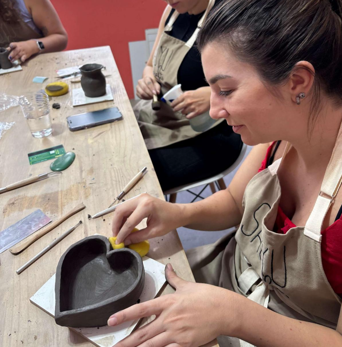 Mulher passando esponja em pote modelado à mão, em formato de coração, durante oficina de cerâmica artesanal no ateliê Shoyu Crafts.
